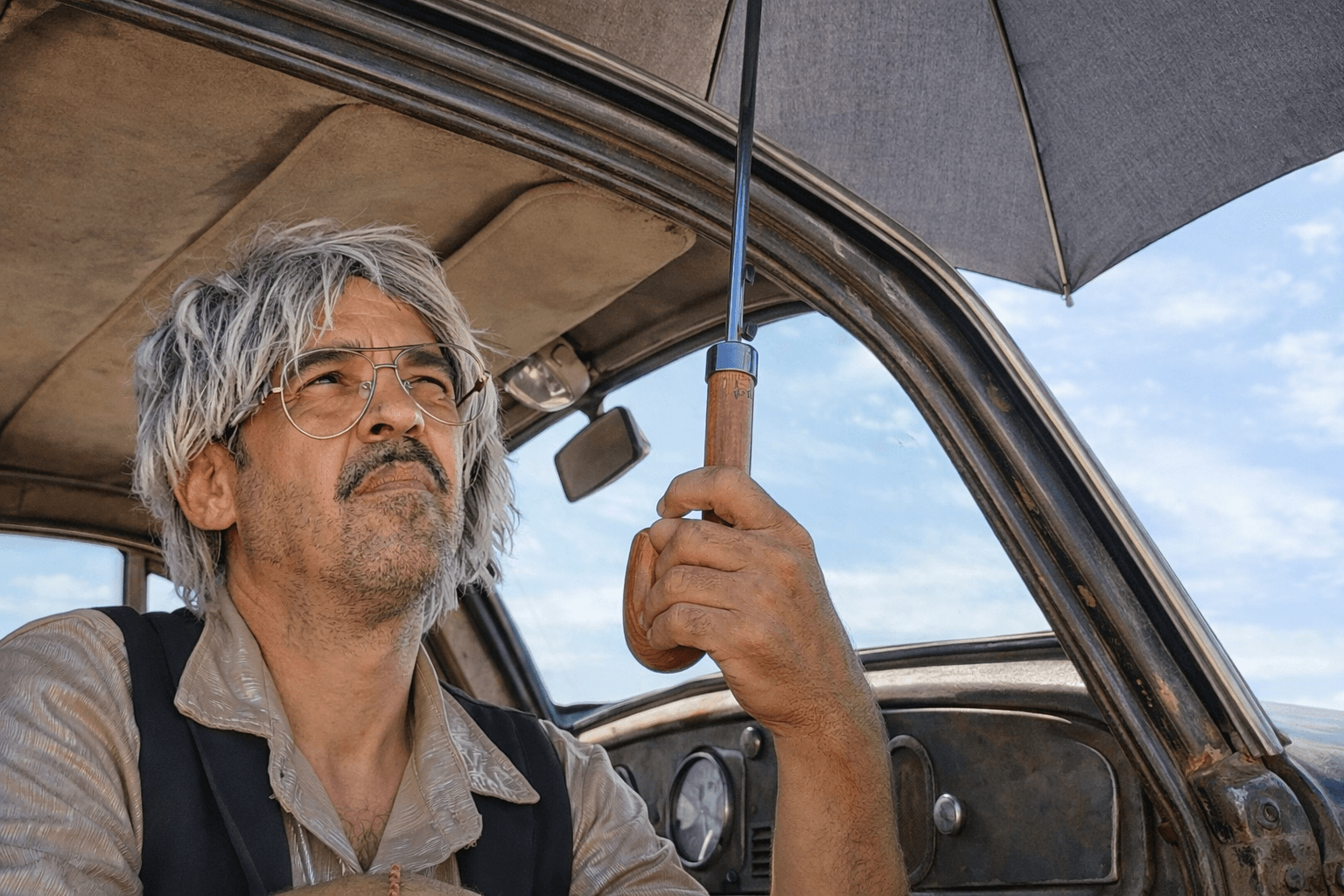 Actor sentado dentro de un coche antiguo mirando al cielo con un paraguas abierto en la mano.