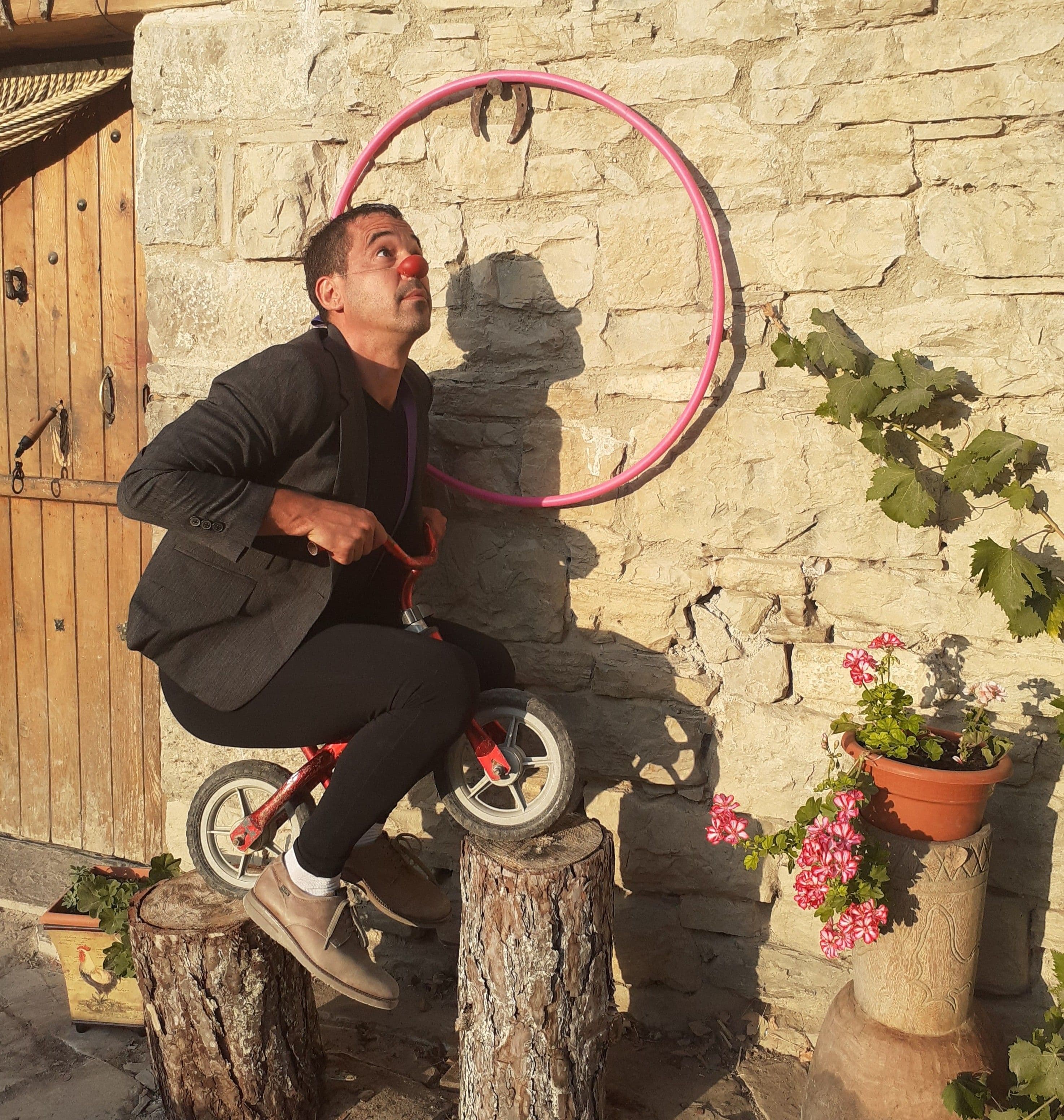 Actor con nariz de payaso haciendo equilibrio en una bicicleta diminuta sobre dos troncos frente a un muro de piedra.