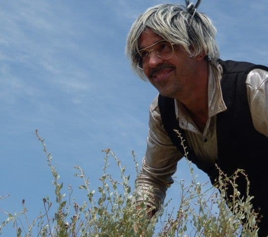 Actor de OseteDelirios caracterizado con peluca y gafas, asomándose entre la hierba en un entorno natural.