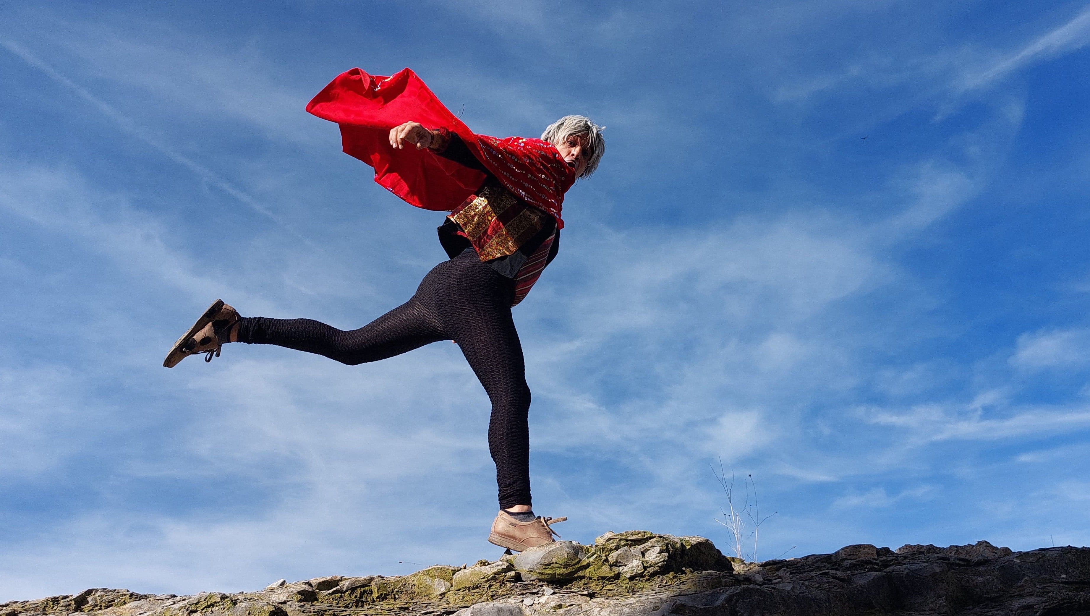 ctor dando un salto sobre unas rocas con una capa roja ondeando al viento bajo un cielo azul.