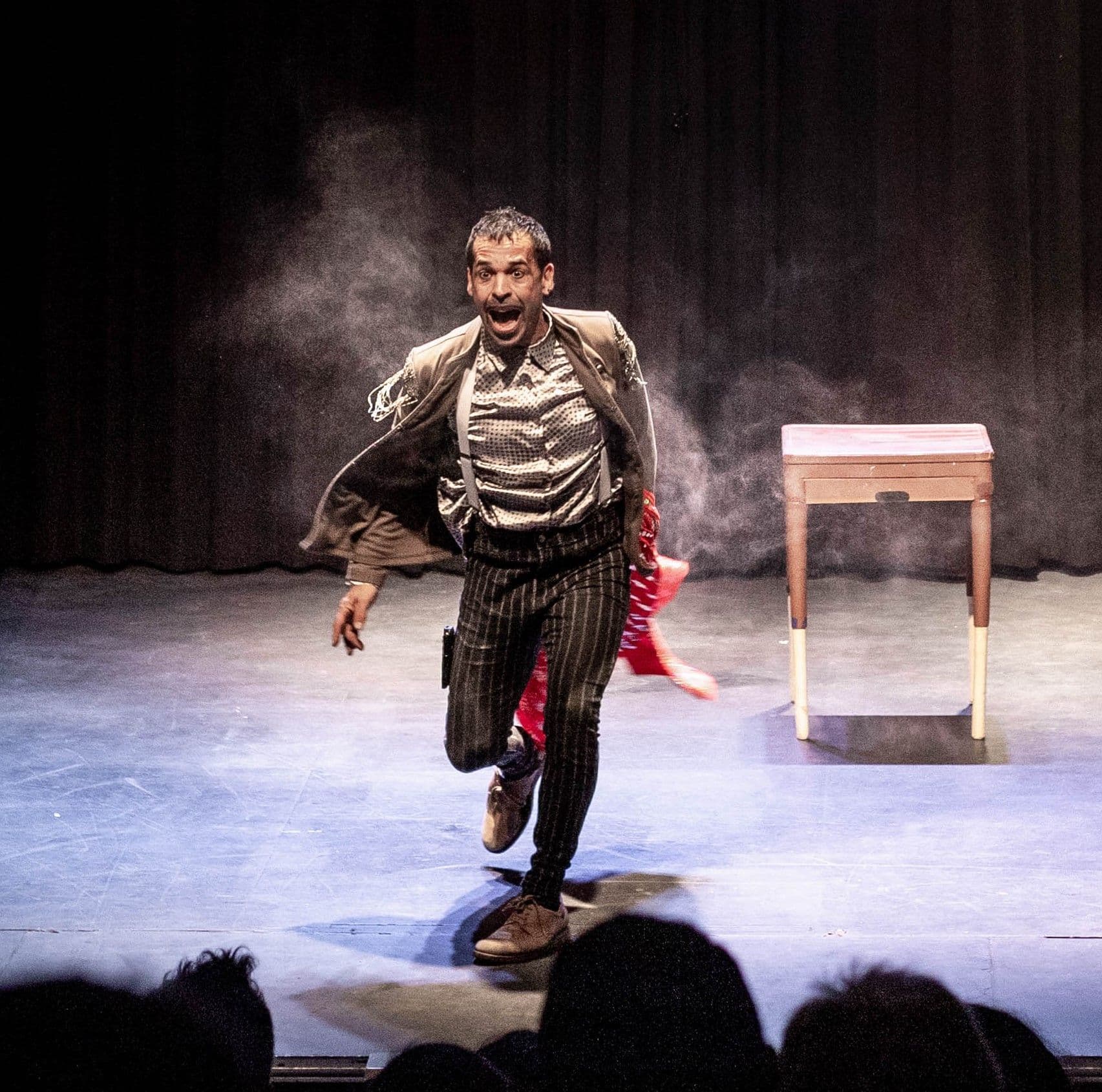 Actor avanzando con intensidad hacia el público en una escena teatral con humo